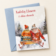 Cerdyn Nadolig i’r Ddau Ohonoch – Sled / Welsh Christmas card with two people on a sledge in the snow, lying flat beside a soft blue envelope.
