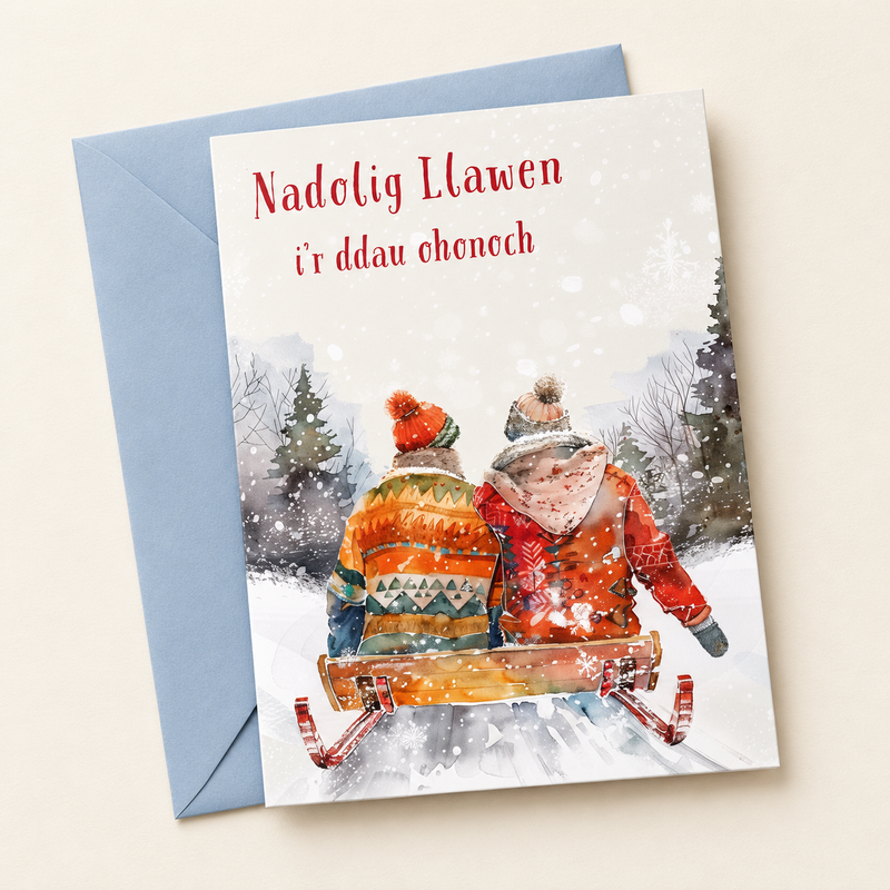 Cerdyn Nadolig i’r Ddau Ohonoch – Sled / Welsh Christmas card with two people on a sledge in the snow, lying flat beside a soft blue envelope.