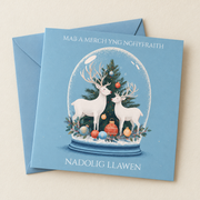 Cerdyn Nadolig Mab a Merch yng Nghyfraith – Ceirw / Welsh Christmas card with white deer and a Christmas tree in a glass dome, lying flat beside a soft blue envelope.