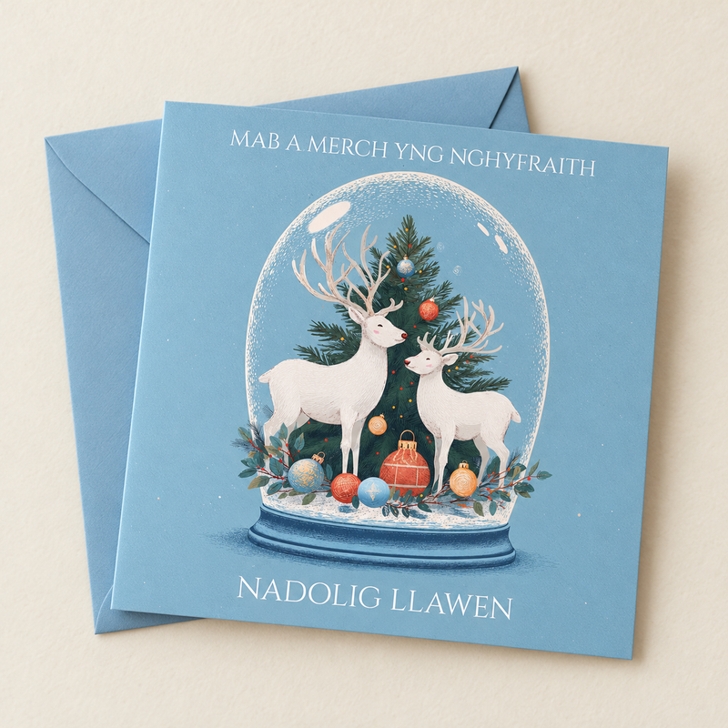 Cerdyn Nadolig Mab a Merch yng Nghyfraith – Ceirw / Welsh Christmas card with white deer and a Christmas tree in a glass dome, lying flat beside a soft blue envelope.