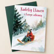Cerdyn Nadolig Mamgu Arbennig – Sled / Welsh Christmas card with a woman sledging in the snow, lying flat beside a deep green envelope.