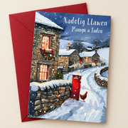 Cerdyn Nadolig Mamgu a Tadcu – Pentref / Welsh Christmas card with a village and a red postbox in the snow, lying flat beside a deep red envelope.