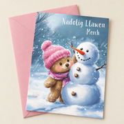 Cerdyn Nadolig Merch – Tedi / Welsh Christmas card with a teddy in a pink hat and a snowman, lying flat beside a soft pink envelope.