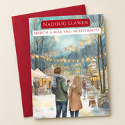 Cerdyn Nadolig Merch a Mab yng Nghyfraith – Marchnad / Welsh Christmas card with two people at a Christmas market, lying flat beside a deep red envelope.