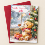 Cerdyn Nadolig Anti – Tedi / Welsh Christmas card with a teddy beside a Christmas tree and window, lying flat beside a deep red envelope.
