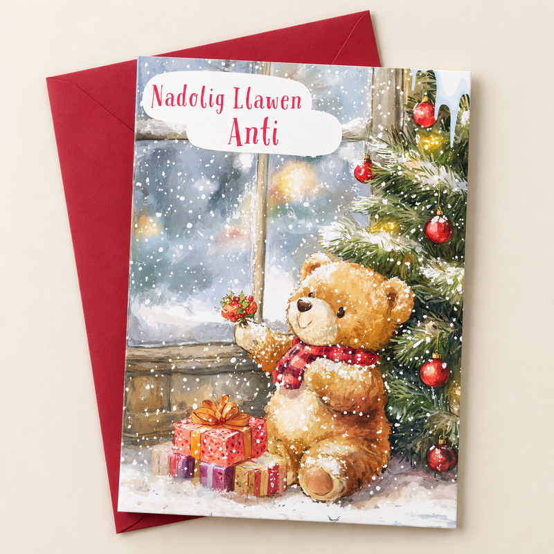 Cerdyn Nadolig Anti – Tedi / Welsh Christmas card with a teddy beside a Christmas tree and window, lying flat beside a deep red envelope.