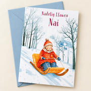 Cerdyn Nadolig Nai – Sled / Welsh Christmas card with a child sledging in the snow, lying flat beside a soft blue envelope.