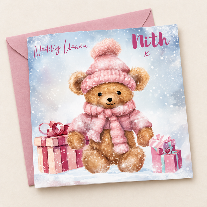 Cerdyn Nadolig Nith – Tedi Pinc / Welsh Christmas card with a teddy in a pink hat and scarf with presents, lying flat beside a soft pink envelope.
