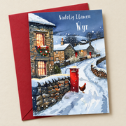 Cerdyn Nadolig Ŵyr – Pentref Eira / Welsh Christmas card with a snowy village and a red postbox, lying flat beside a deep red envelope.
