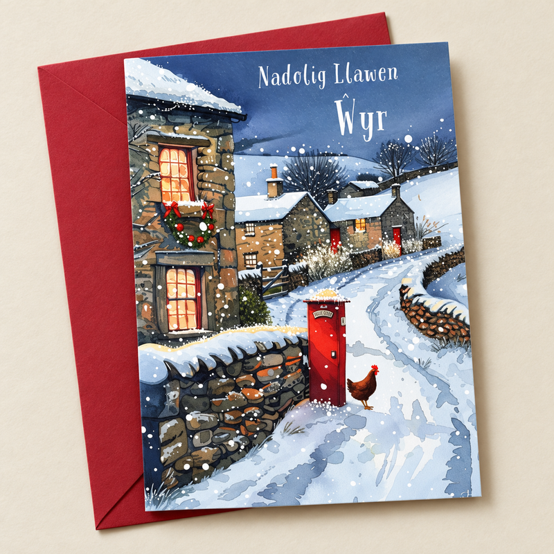 Cerdyn Nadolig Ŵyr – Pentref Eira / Welsh Christmas card with a snowy village and a red postbox, lying flat beside a deep red envelope.
