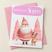 Cerdyn Nadolig Ŵyres – Siôn Corn / Welsh Christmas card with Father Christmas and a small reindeer in a pink scene, lying flat beside a soft pink envelope.